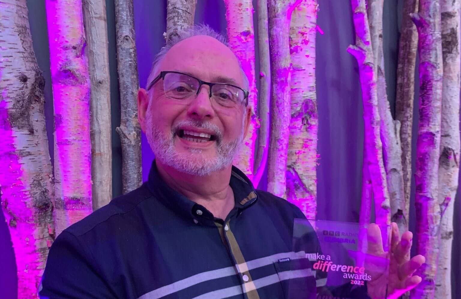 Man holding an award with the text "make a difference awards 2022," standing in front of birch trees and pink lighting. - Home Instead
