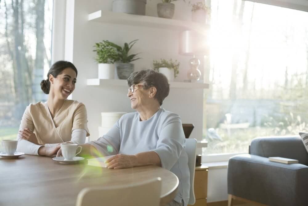 Two women sitting at a table with coffee, smiling at each other in a sunlit room with plants and modern decor. - Home Instead