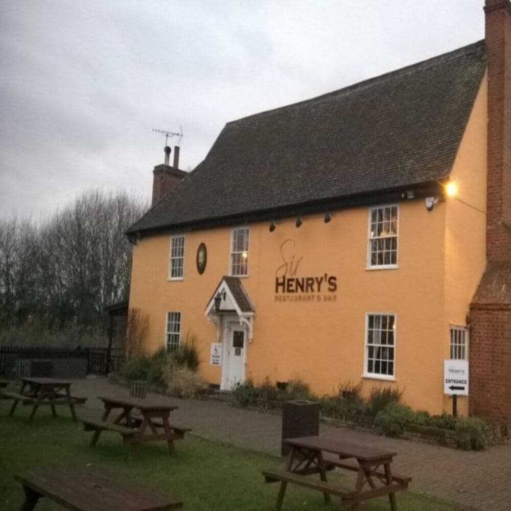 A yellow restaurant and bar named "Henry's" with picnic benches on a grassy area in front, under a cloudy sky. - Home Instead