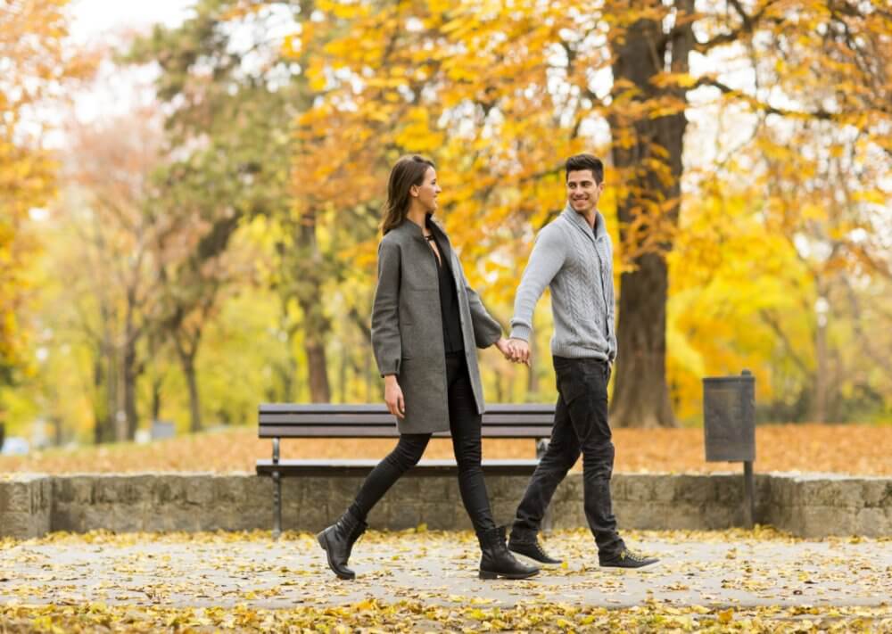 A couple walking hand in hand through a park with autumn foliage in the background. - Home Instead
