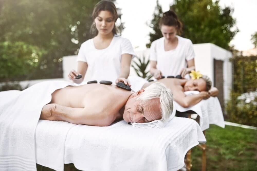 Two people receiving hot stone massages outdoors by two massage therapists in white uniforms. - Home Instead