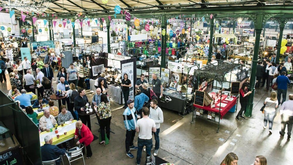 A bustling indoor market with people browsing various stalls and socializing under decorative banners and lights. - Home Instead