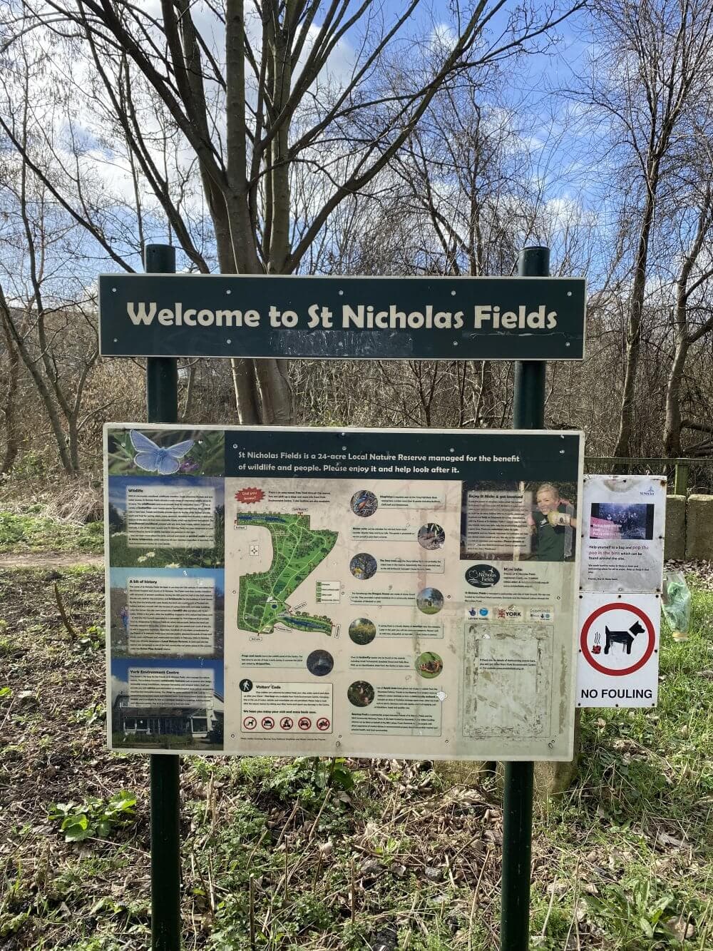 Sign reading "Welcome to St Nicholas Fields" with information about the nature reserve, set in a wooded area. - Home Instead