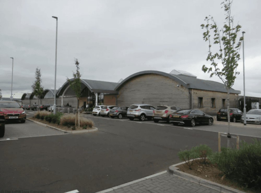 Parking lot in front of a modern industrial building with curved roofs, several parked cars, and some small trees. - Home Instead