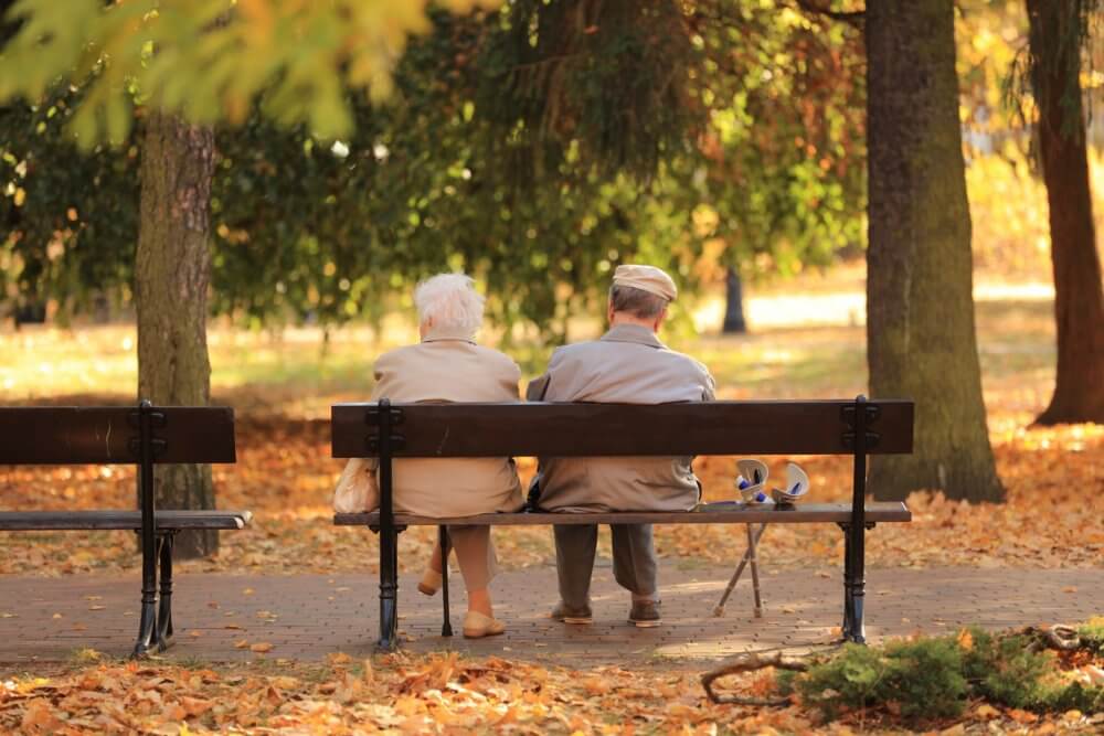 An elderly couple sits on a park bench surrounded by autumn leaves, facing away and enjoying a peaceful moment. - Home Instead