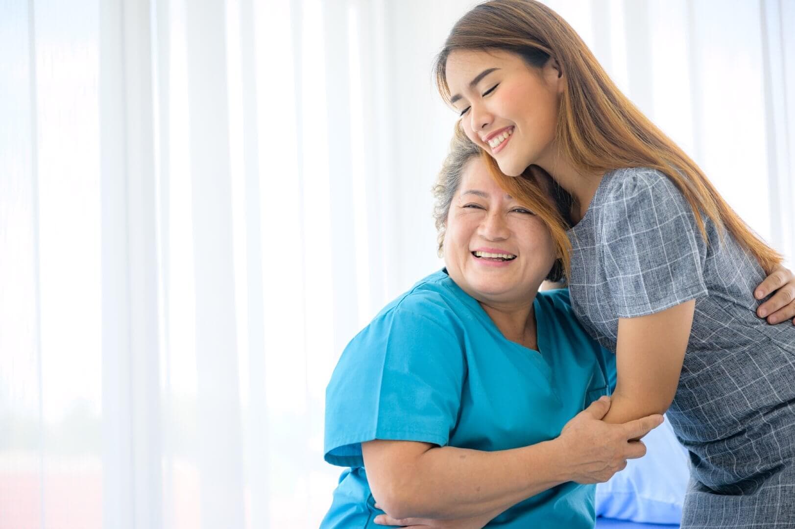 A young woman warmly embraces an older woman in teal scrubs, both smiling brightly in a well-lit room. - Home Instead Bournemouth & Christchurch