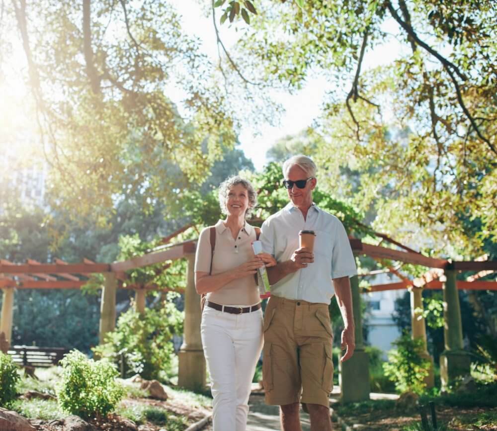 An elderly couple walking and smiling in a sunlit park, holding drinks under leafy trees. - Home Instead