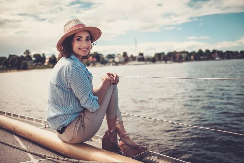 Woman in a hat sitting on a boat, smiling with a lake and trees in the background on a sunny day. - Home Instead Bournemouth & Christchurch