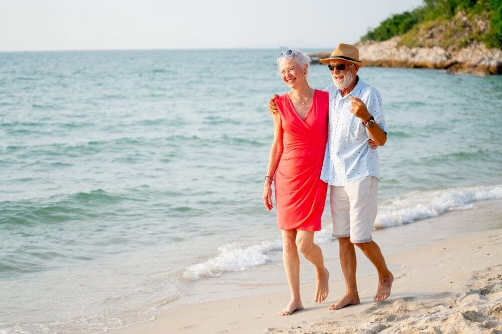 An elderly couple walks arm in arm on a beach. The woman is in a red dress, and the man wears a hat and white shirt. - Home Instead Bournemouth & Christchurch