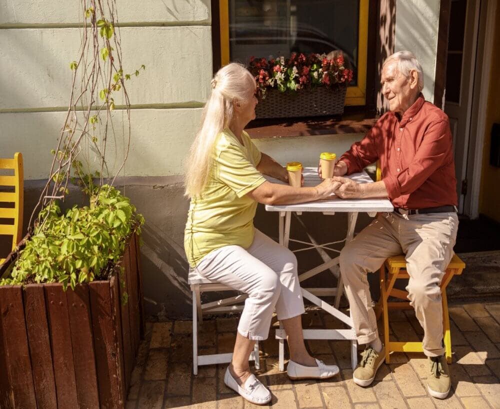 Elderly couple sitting at an outdoor café table, smiling and holding hands, with coffee cups in front of them. - Home Instead Bournemouth & Christchurch