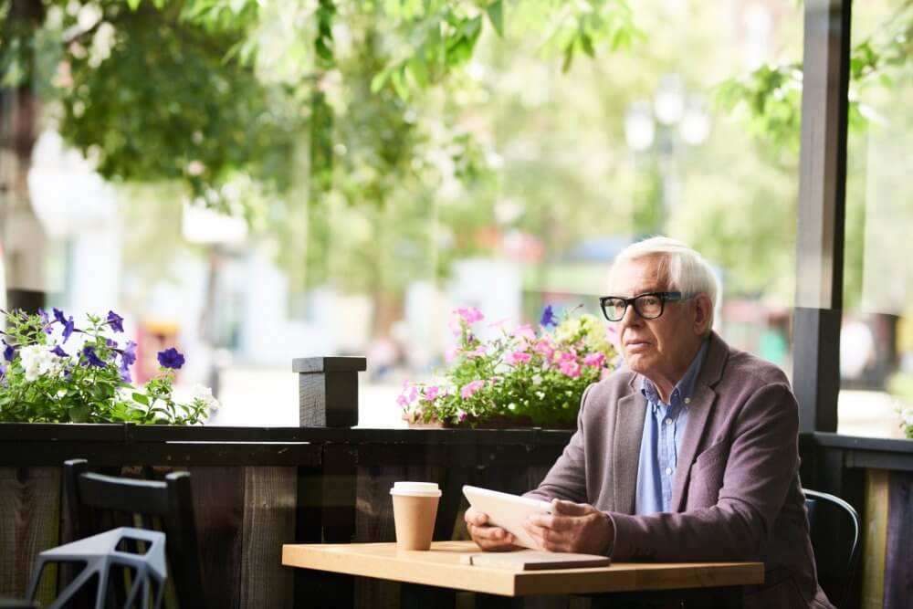Elderly man with glasses reading a tablet at an outdoor café table with flowers and greenery in the background. - Home Instead Bournemouth & Christchurch