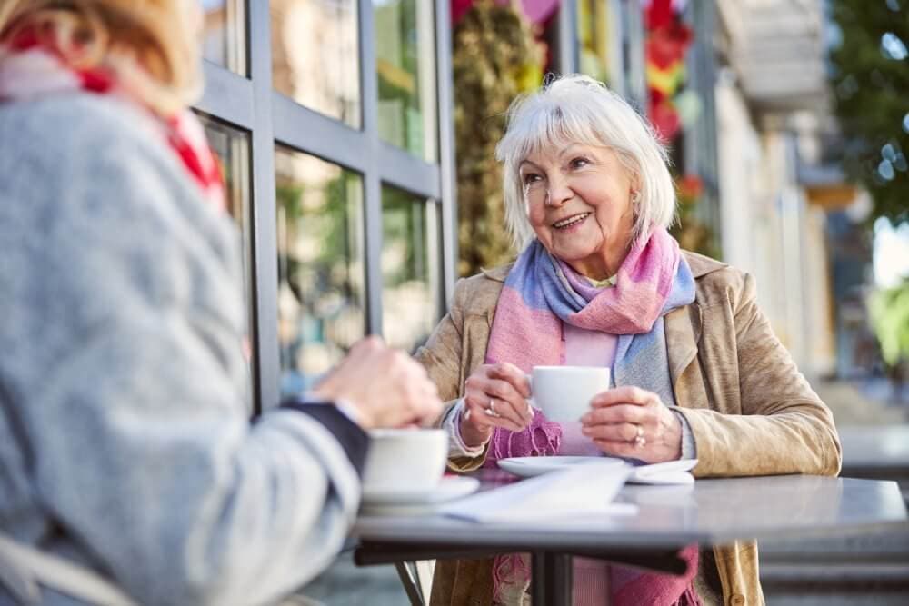 Two elderly women enjoying coffee at an outdoor café, smiling and engaged in conversation on a sunny day. - Home Instead