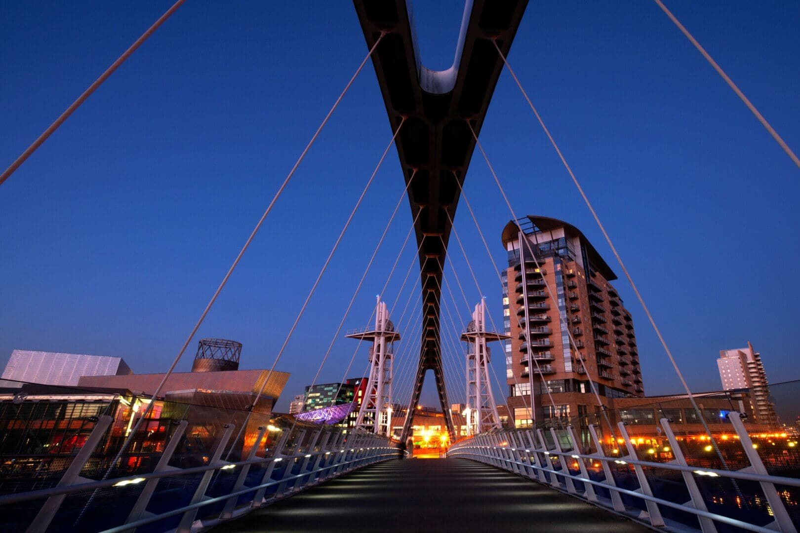 A modern pedestrian bridge illuminated at twilight, with city buildings and a clear blue sky in the background. - Home Instead
