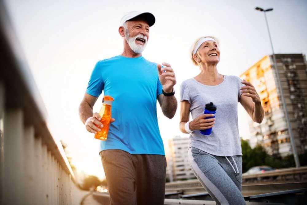 A senior man and woman enjoy a jogging session while holding water bottles, on an urban path with buildings in the background. - Home Instead