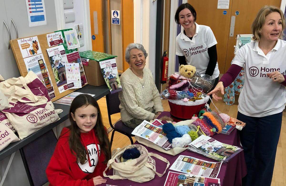 Four people at a Home Instead event, showcasing crafts and flyers on a table, with knitting materials and bags. - Home Instead