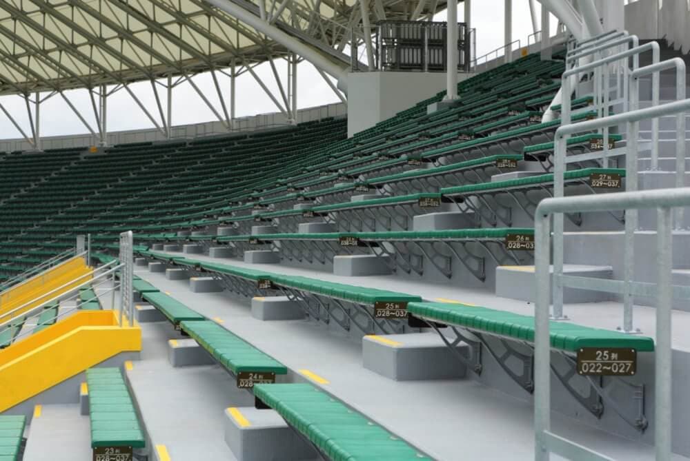 Stadium seating with green benches and yellow stair railings under a large, partially covered roof. - Home Instead