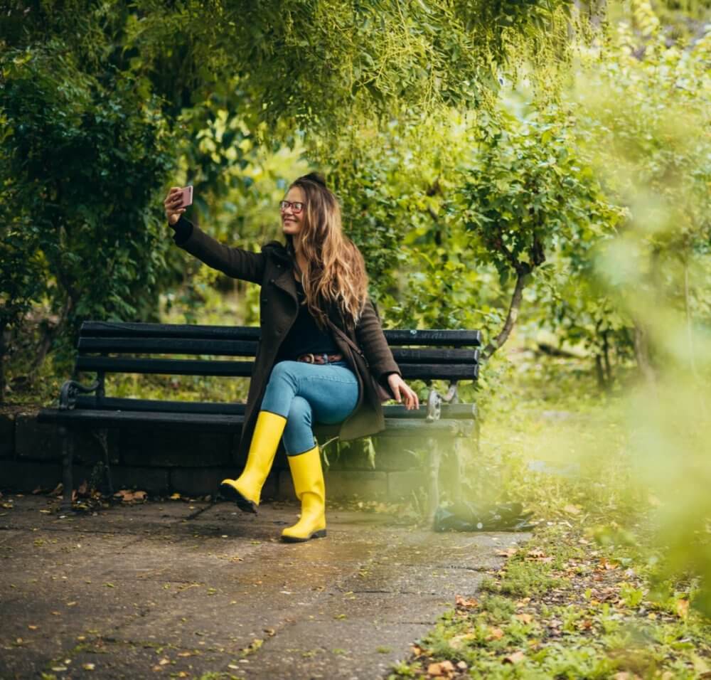A woman in a coat and yellow rain boots sits on a bench, taking a selfie in a lush garden. - Home Instead Bournemouth & Christchurch