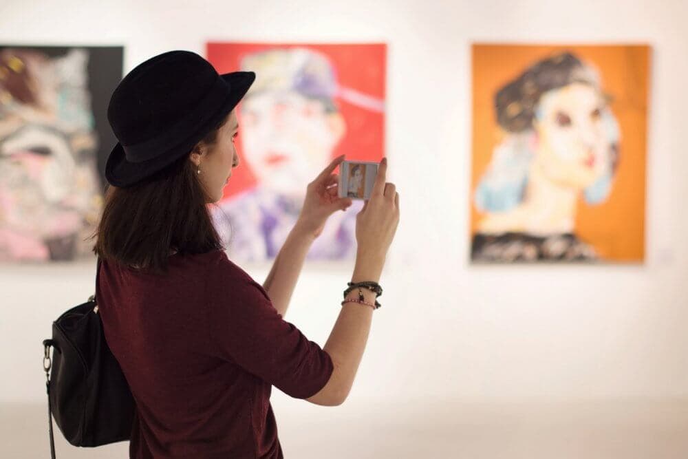 A woman in a hat taking a photo with her phone of abstract art in a gallery. - Home Instead Bournemouth & Christchurch