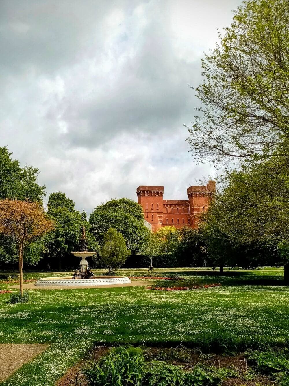 A view of a red brick castle with round towers, surrounded by lush green trees and a garden with a central fountain. - Home Instead