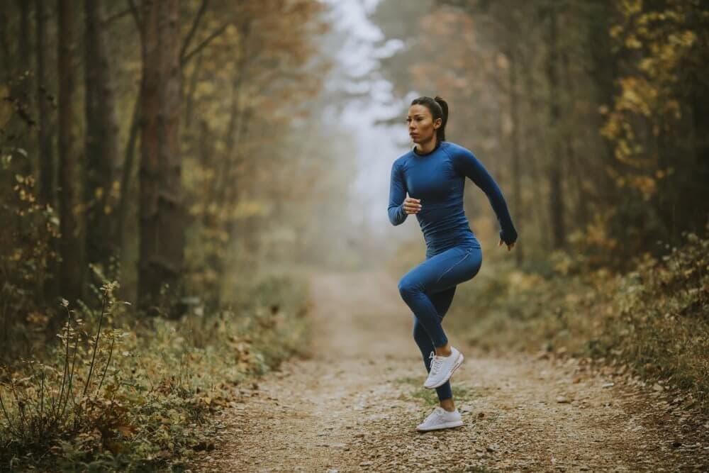 Person in blue athletic wear jogging on a forest path surrounded by tall trees and autumn foliage. - Home Instead