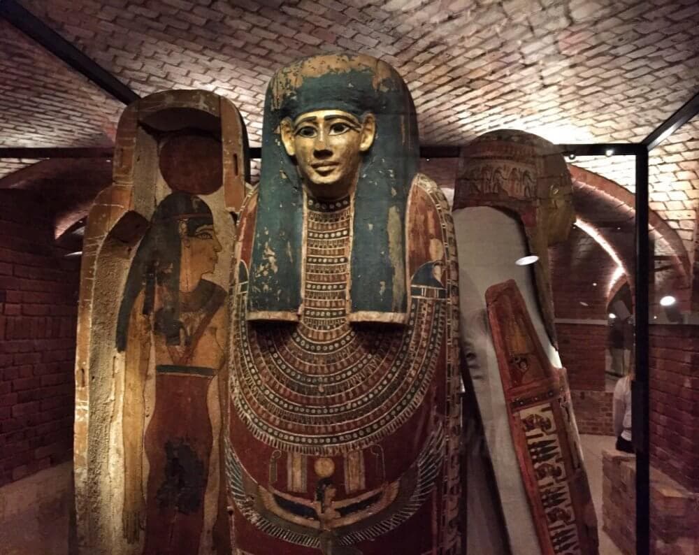 Three ancient Egyptian sarcophagi displayed in a museum exhibit under arched brick ceilings. - Home Instead
