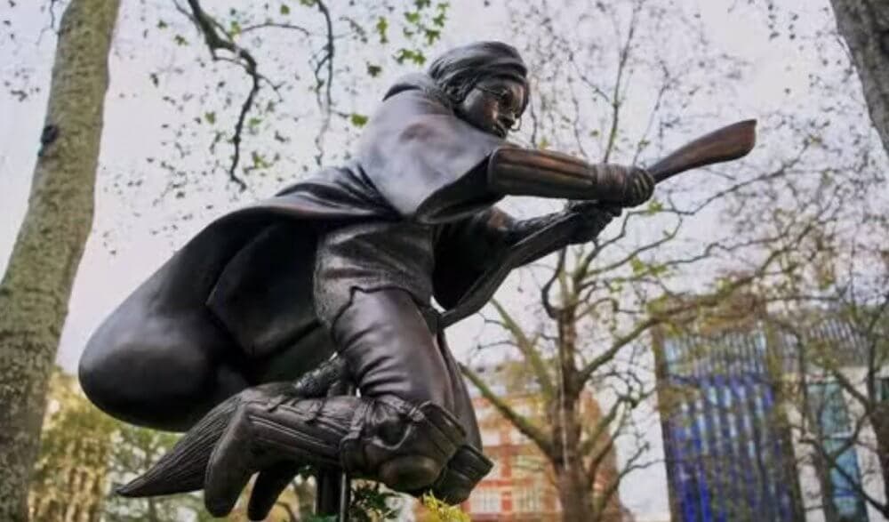 Statue of a figure flying on a broomstick in a forested area, with buildings visible in the background. - Home Instead