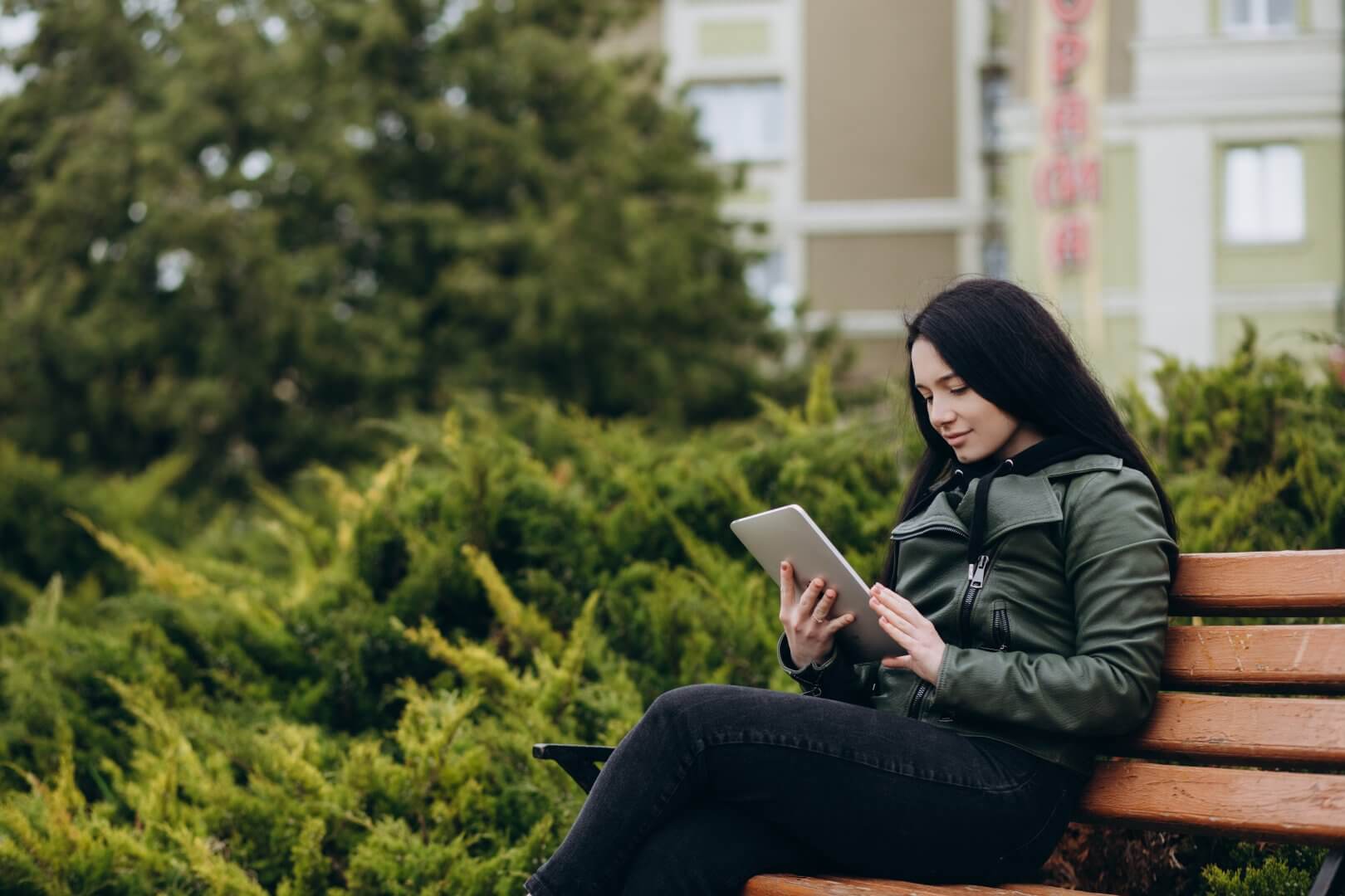 A person sits on a bench in a park, using a tablet while surrounded by greenery and buildings in the background. - Home Instead