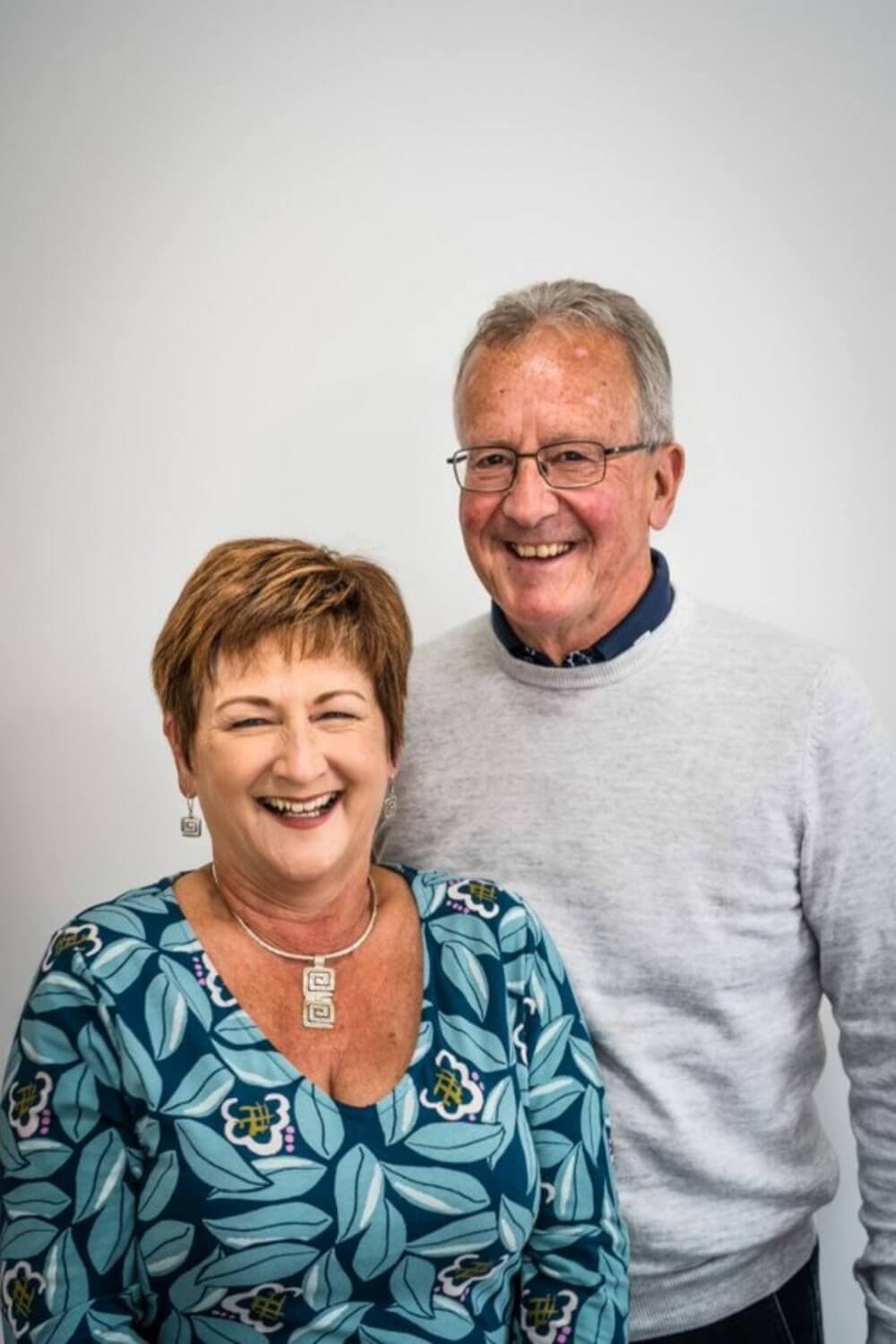 Two smiling people, one in a patterned shirt and the other in a light sweater, standing in front of a plain background. - Home Instead