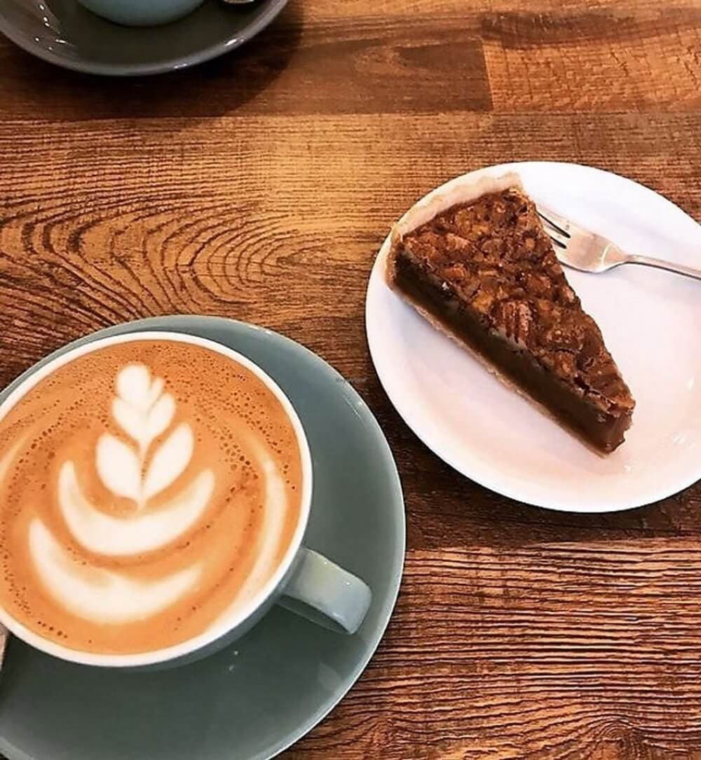 A cup of latte with leaf latte art and a slice of pecan pie on a plate sit on a wooden table. - Home Instead