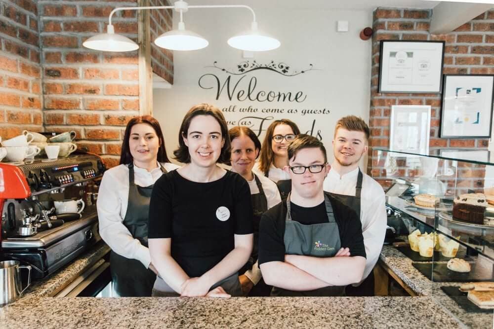 A group of six people, three men and three women, smiling behind a cafe counter with cakes. - Home Instead