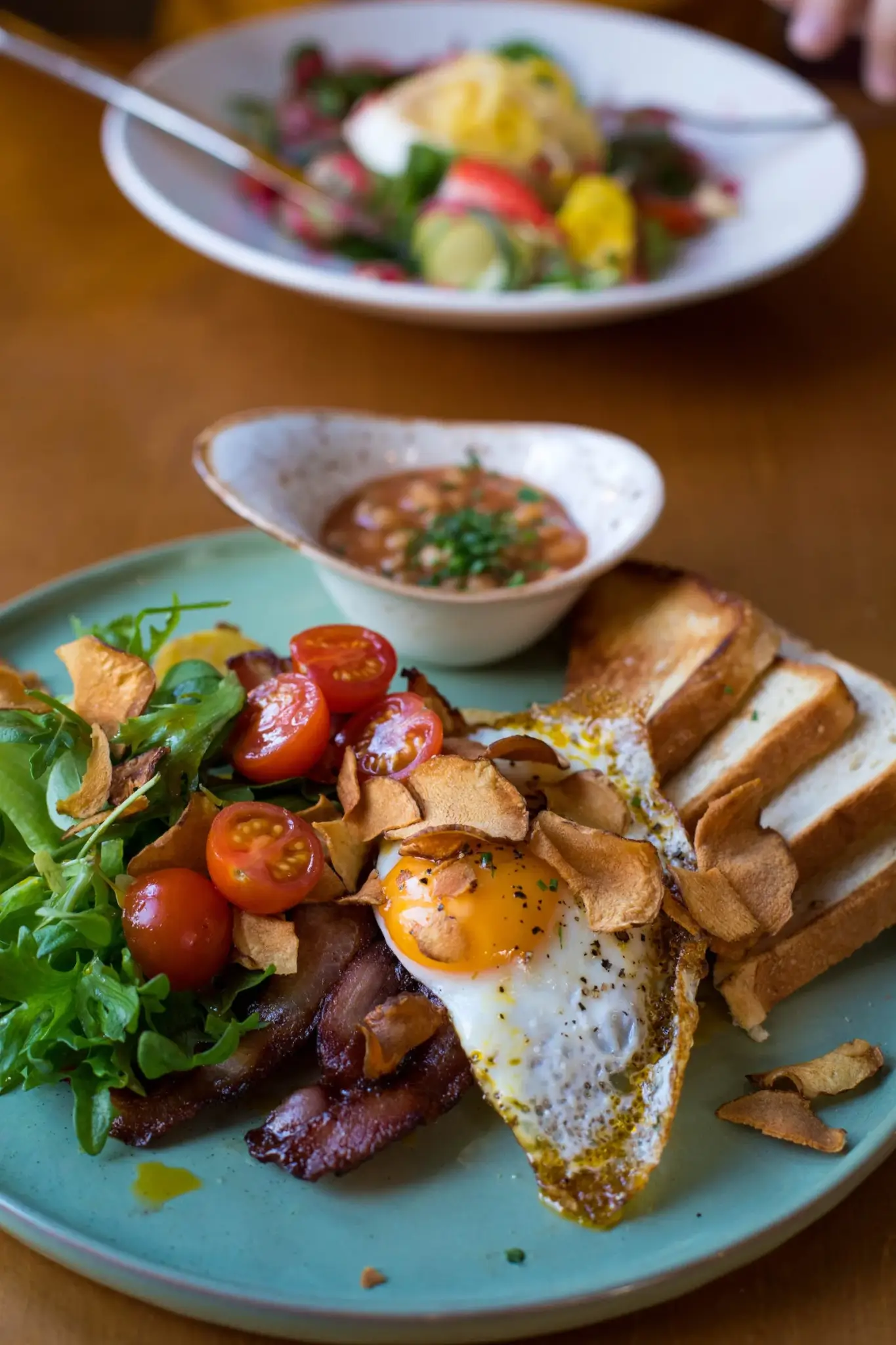 A plate with fried egg, cherry tomatoes, greens, toast, and a small dish of salsa, with a salad in the background. - Home Instead