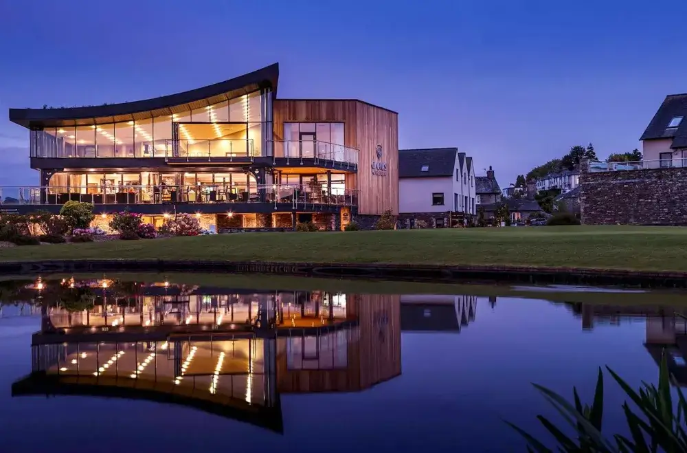 Modern building with large windows and a curved roof, reflecting on a calm pond, during twilight. - Home Instead