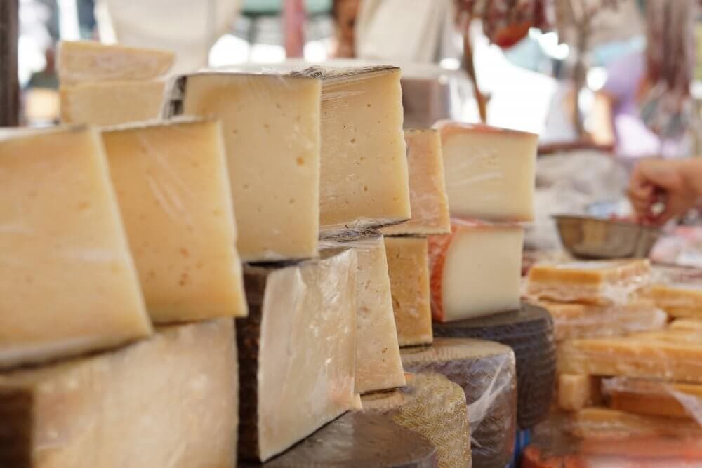 Stacks of assorted square and triangular cheese blocks on display at a market stall. - Home Instead