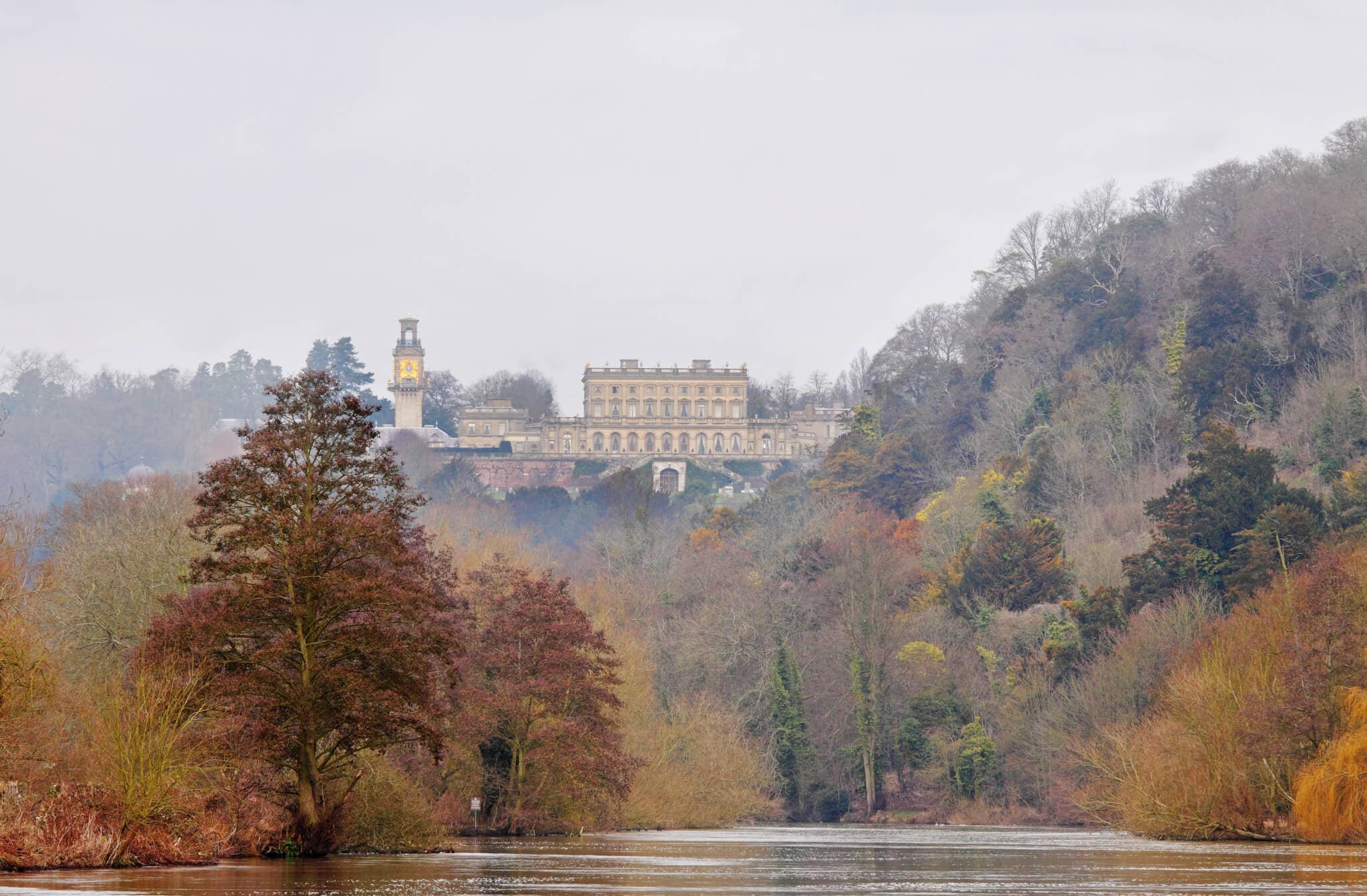 A stately home on a hill, surrounded by trees in autumn colors, with a river in the foreground. - Home Instead