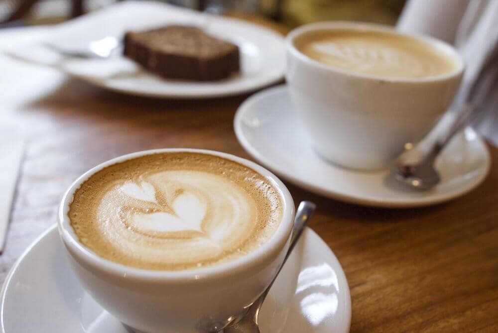 Two cups of latte with latte art on a wooden table, accompanied by a plate of bread in the background. - Home Instead