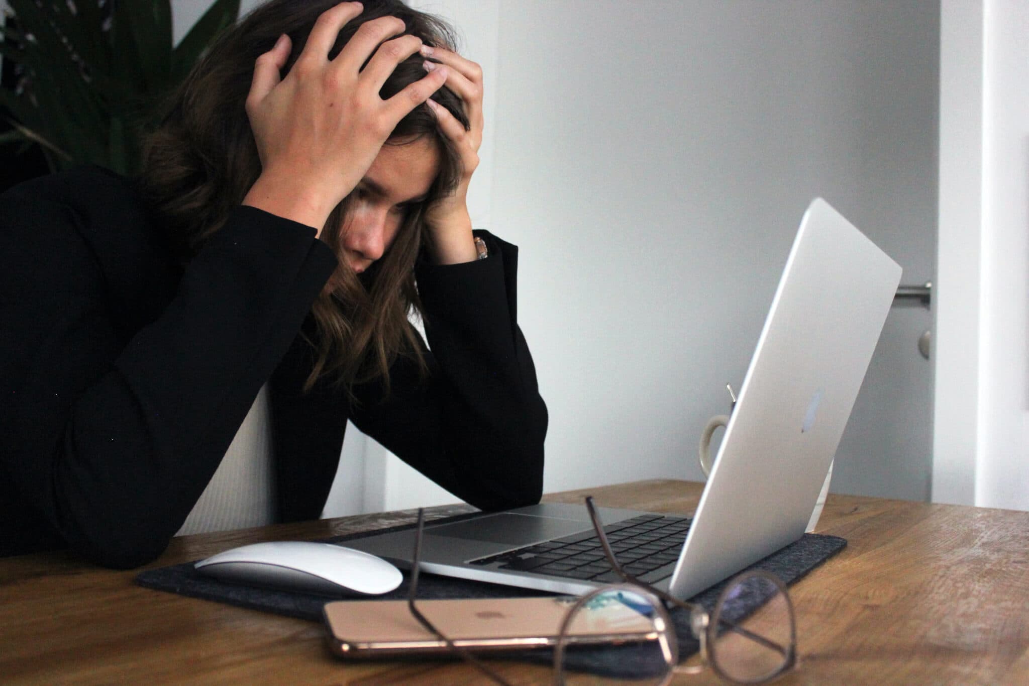 Person holding their head with hands, sitting in front of a laptop on a desk with glasses and a phone. - Home Instead