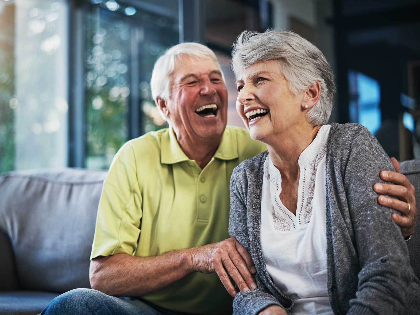 Elderly couple sitting on a couch, laughing and embracing each other in a bright living room. - Home Instead