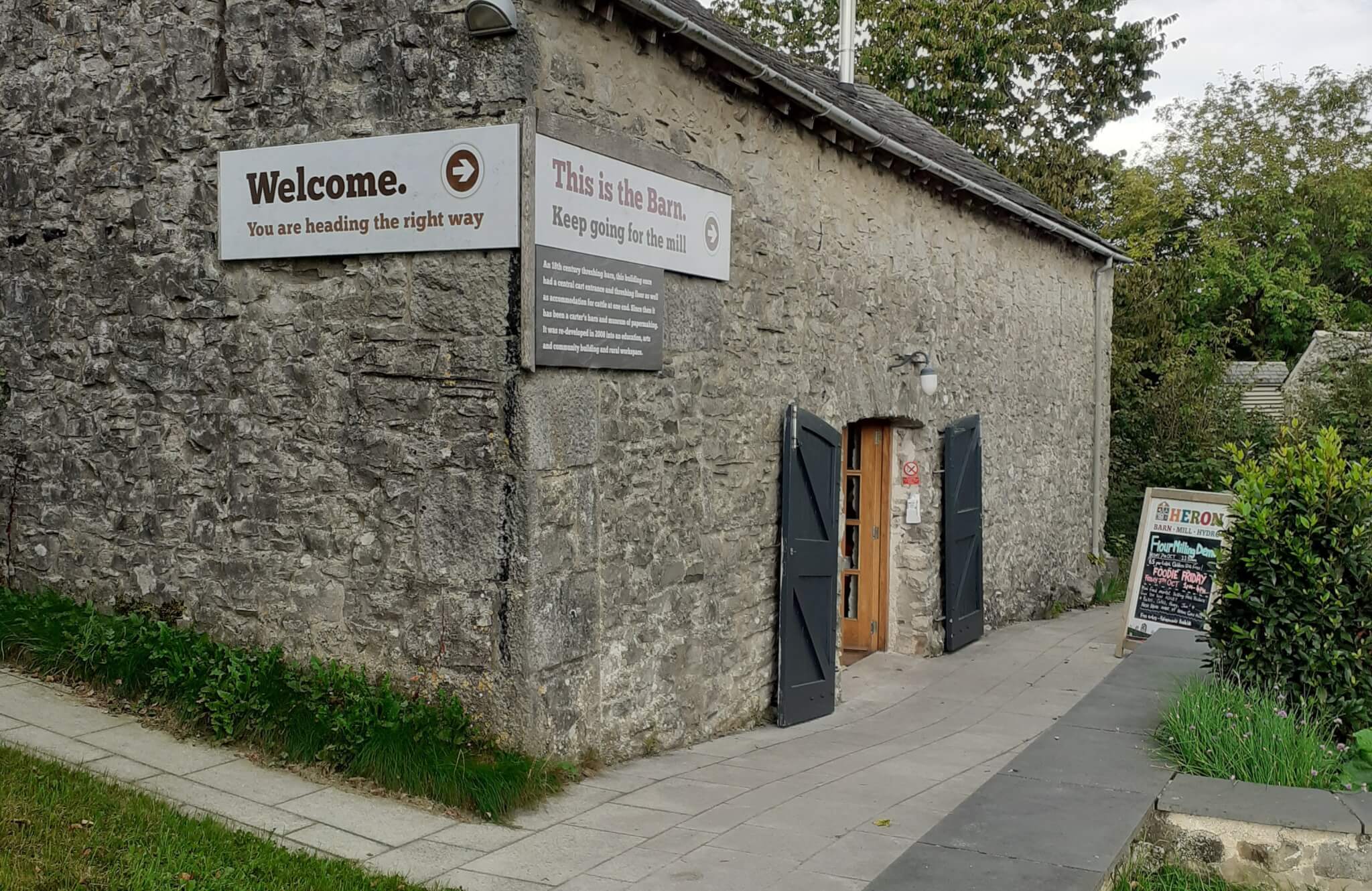 Stone building with open doors and welcoming signs that say "This is the Barn. Keep going for the fun!" and "Welcome. - Home Instead