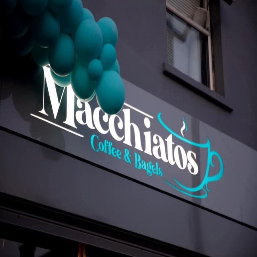 Sign for "Macchiatos Coffee & Bagels" on a dark building, with teal balloons above it. - Home Instead