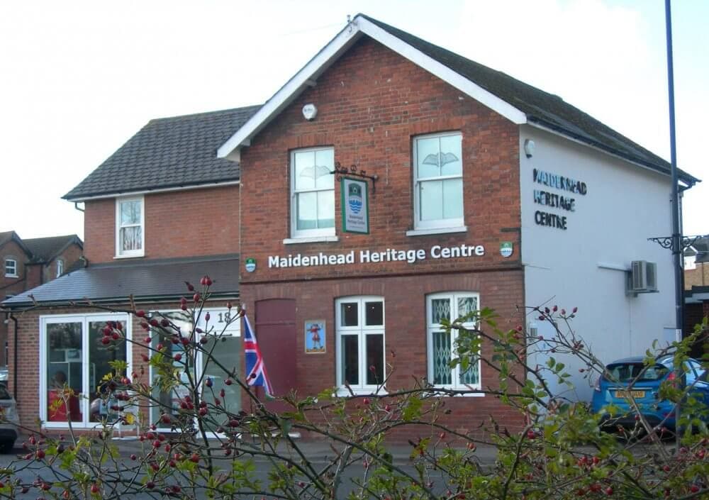 A two-story brick building with a sign for Maidenhead Heritage Centre, surrounded by trees and bushes. - Home Instead
