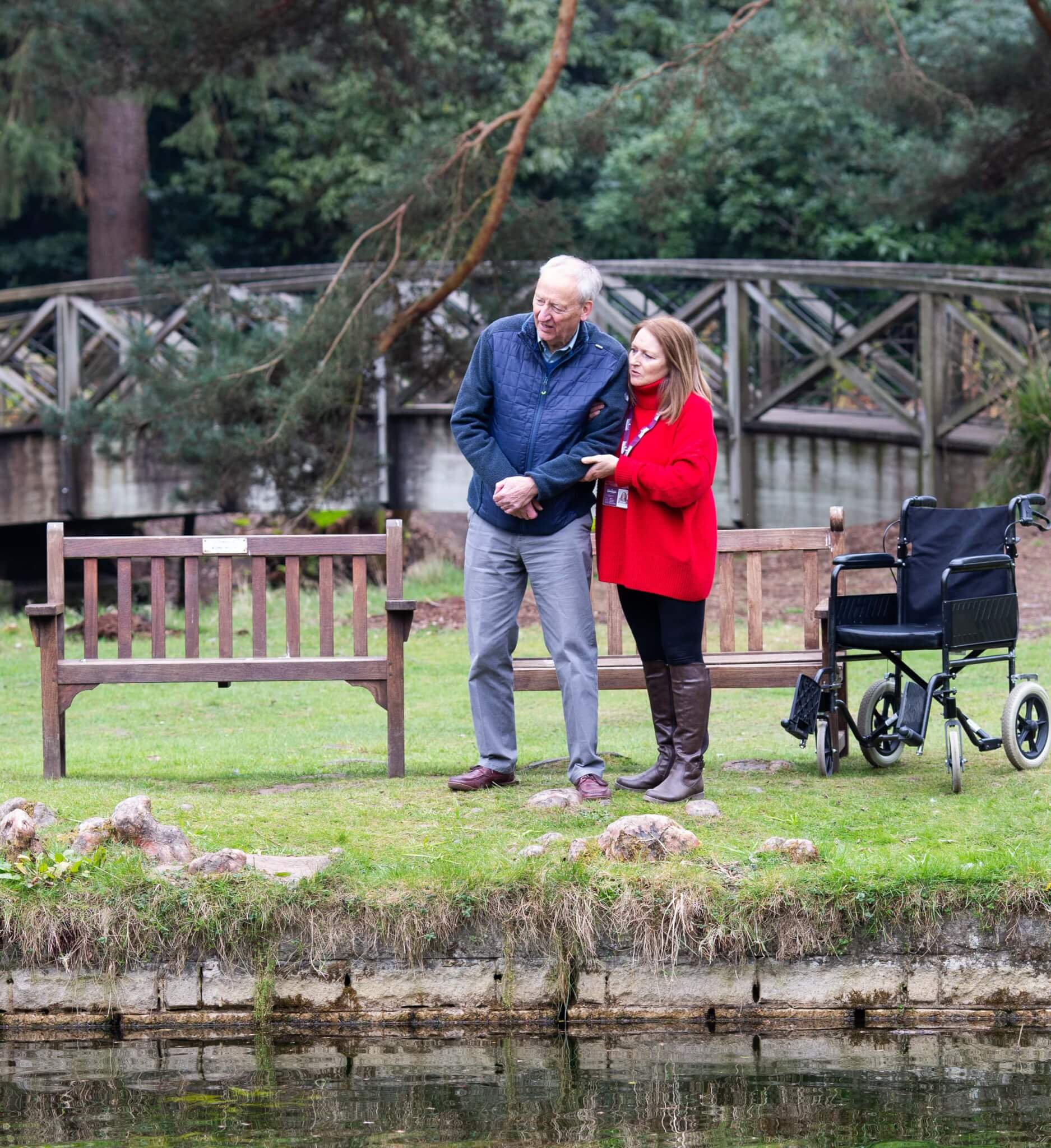 An elderly man and a woman stand by a pond, with an empty wheelchair nearby and a wooden bridge in the background. - Home Instead