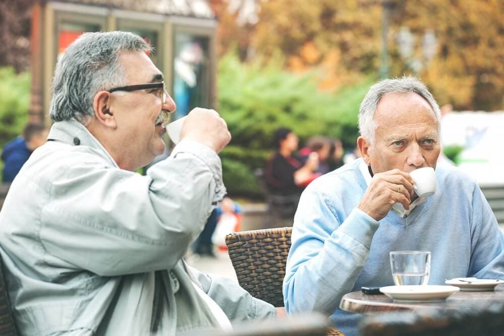Two elderly men sit at an outdoor café enjoying coffee, one laughs while the other takes a sip from a white cup. - Home Instead