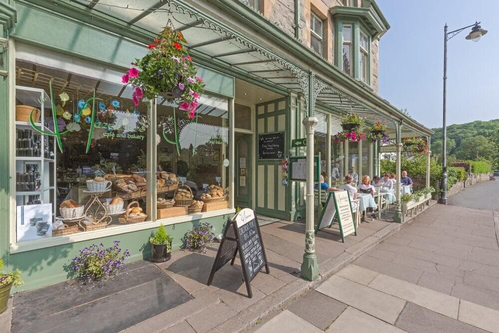 A quaint bakery with a green facade, outdoor seating, hanging flower baskets, and a chalkboard sign on a sunny day. - Home Instead