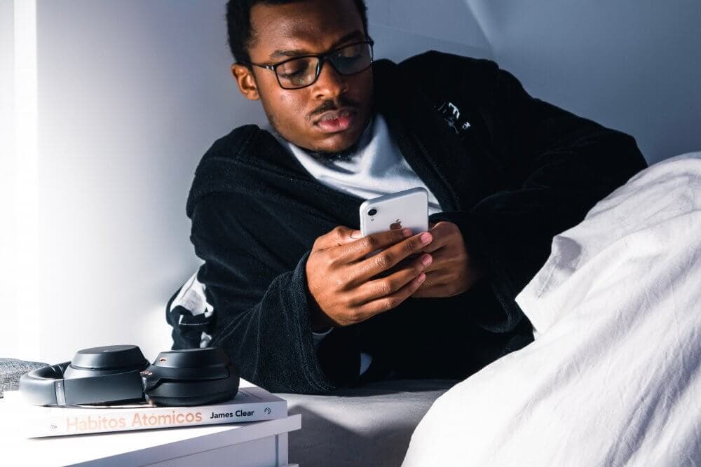 Person in a dark robe looking at a phone in bed, with a book and headphones on the bedside table. - Home Instead