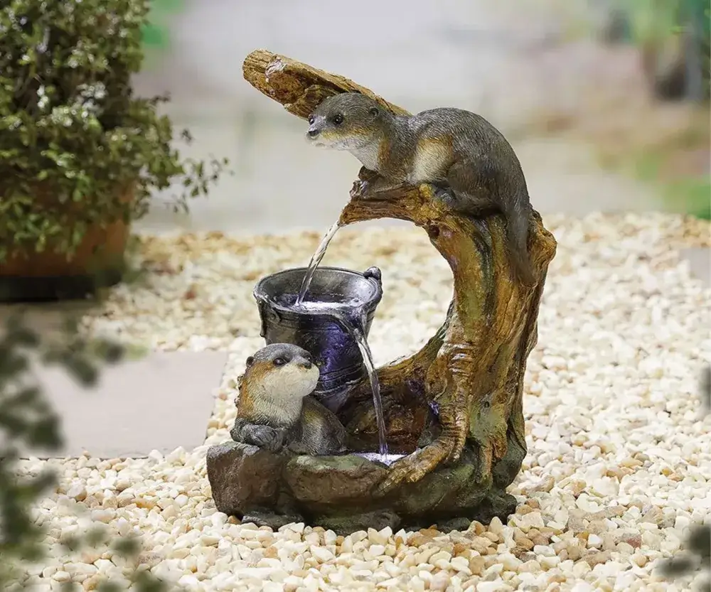 Two otter sculptures with a small water fountain feature, surrounded by pebbles and greenery. - Home Instead