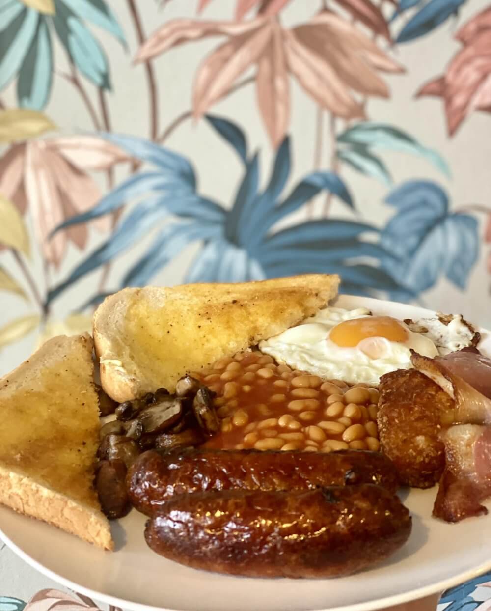 Full English breakfast with sausages, bacon, baked beans, mushrooms, fried egg, hash browns, and toast on a white plate. - Home Instead