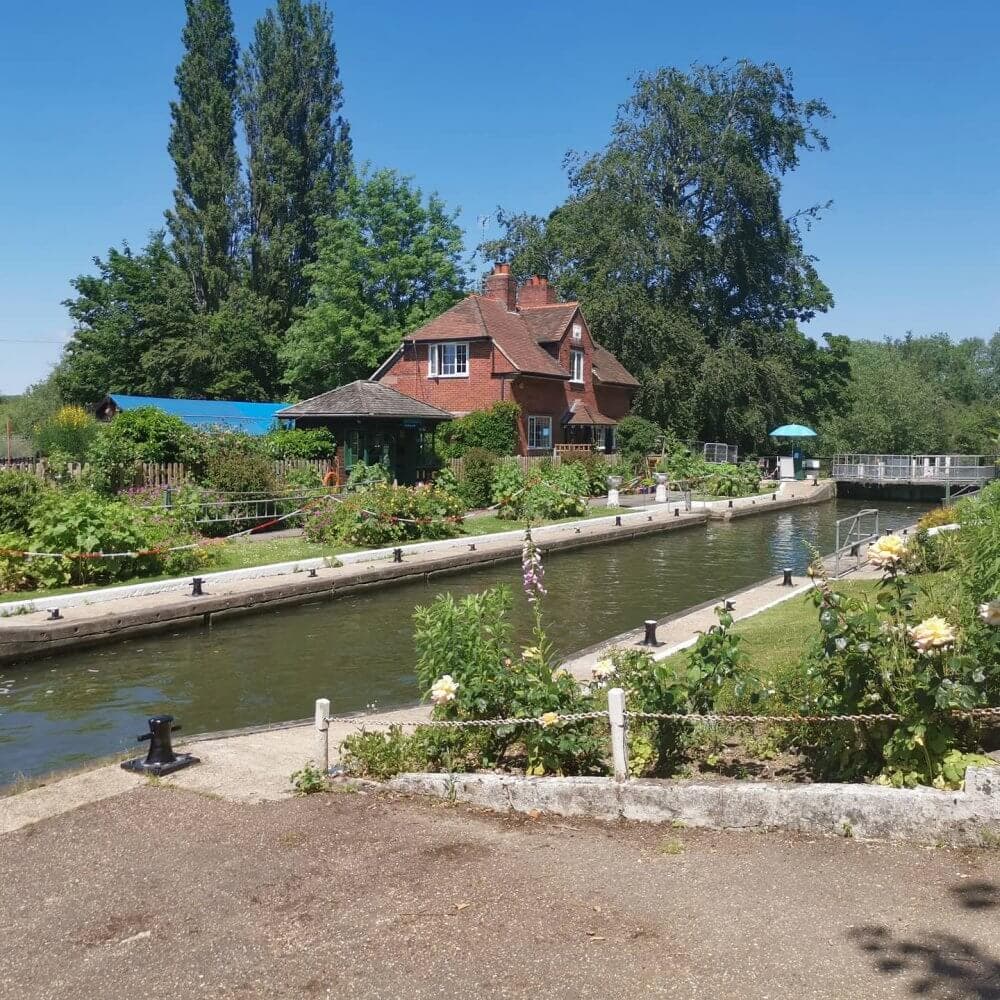 A red-brick house sits beside a peaceful canal surrounded by greenery and flowers under a clear blue sky. - Home Instead
