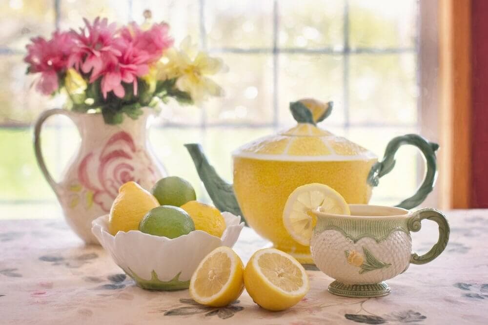 A decorative lemon-themed tea set with flowers, lemons, limes, and a window in the background on a floral tablecloth. - Home Instead