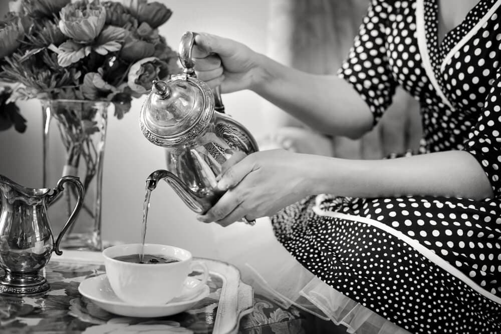 A person in a polka dot dress pours tea from a teapot into a cup next to a bouquet of flowers. - Home Instead