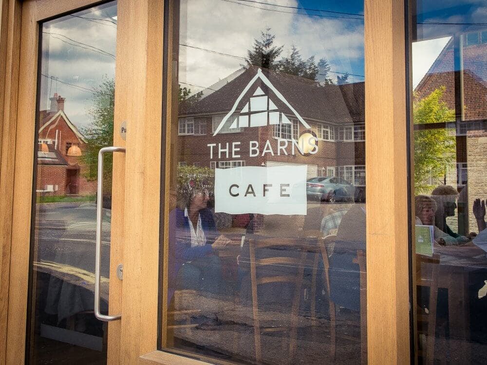 Glass door of "The Barns Cafe" reflecting street view with people sitting inside and buildings in the background. - Home Instead